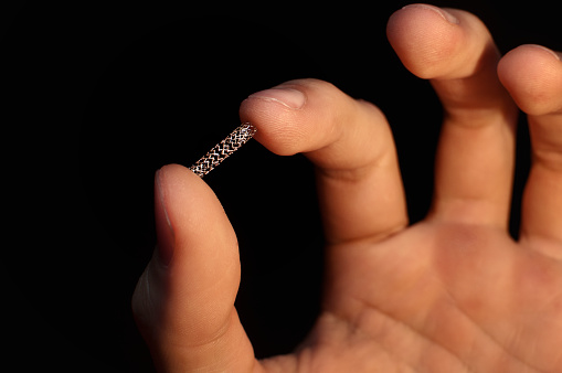 Man's hand holding a stent between forefinger and thumb in front of a clear black background.