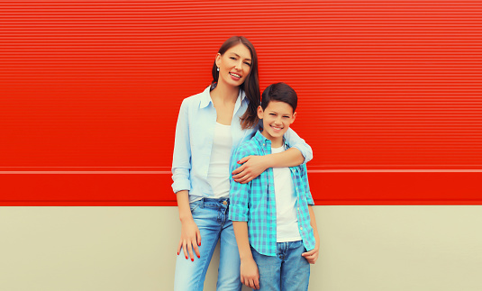 Stylish happy smiling mother with son teenager posing together in checkered shirts, jeans in the city on vivid red background