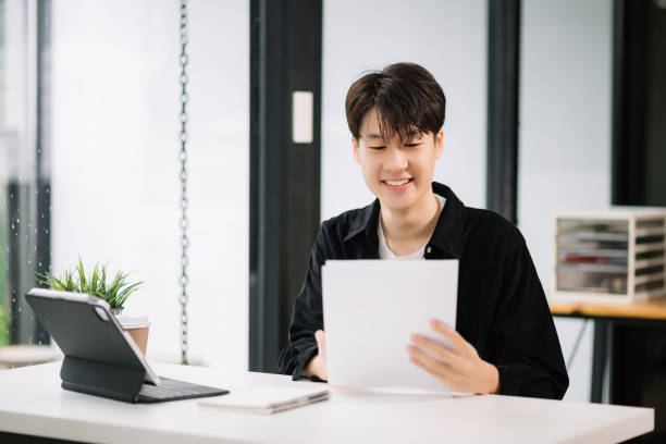 istock Young attractive Asian man smiling thinking planning writing in notebook, tablet and laptop working from home, looking at camera at office 1589156464
