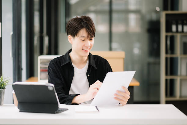 istock Young attractive Asian man smiling thinking planning writing in notebook, tablet and laptop working from home, looking at camera at office 1589156469
