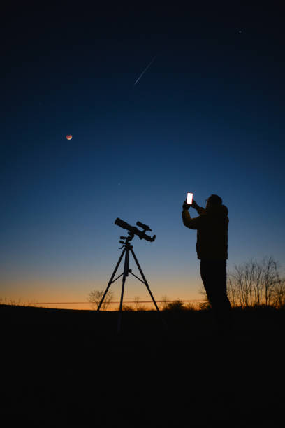 istock Astronomer looking at the starry skies with planets, falling stars and Moon eclipse with a telescope while using modern smartphone. 1589319303