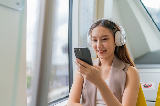 With headphones on, the young Asian businesswoman enjoys interactive social content on her phone during her commute, finding moments of personal entertainment and connection