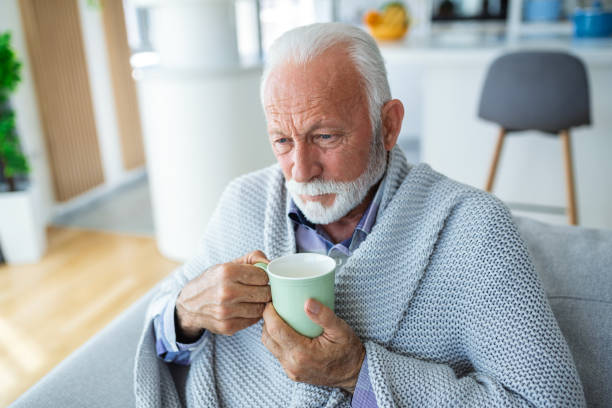istock Senior man suffering from flu drinking tea while sitting wrapped in a blanket on the sofa at home. Sick older man with headache sitting under the blanket in the living room. Man with a cold lying on the sofa holding a mug of hot tea. 1589338947