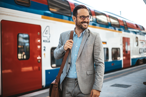 Photo of a smiling businessman at train station.  Confident professional is wearing suit. He is standing at railroad station platform. Businessman at the Train Station