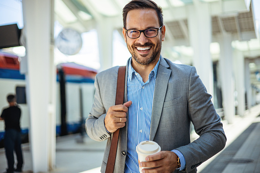 A traveling male business person is at the train station with a reusable travel mug of coffee. A man in a train station commuting to work. Businessman getting in passenger train and going home after working day.