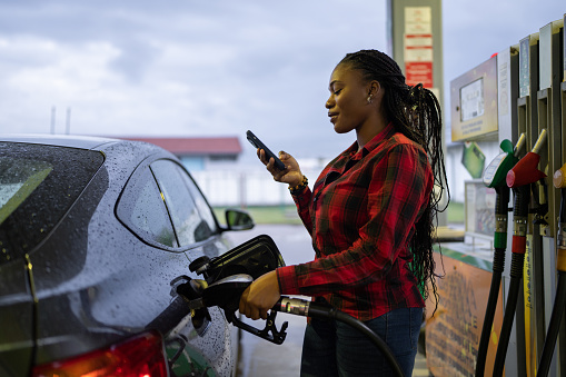 Beautiful black woman pouring gas into her car and using smart phone at the gas station.