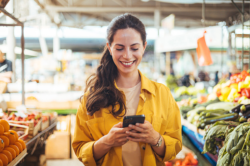 Woman at the farmer's market shopping, sending a text message on her smart phone. Young cheerful woman at the market. Smiling girl decided to cook a delicious and healthy meal