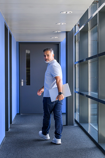 Mature businessman with blank expression and a laptop under his arm walking in a corridor  looking over his shoulder