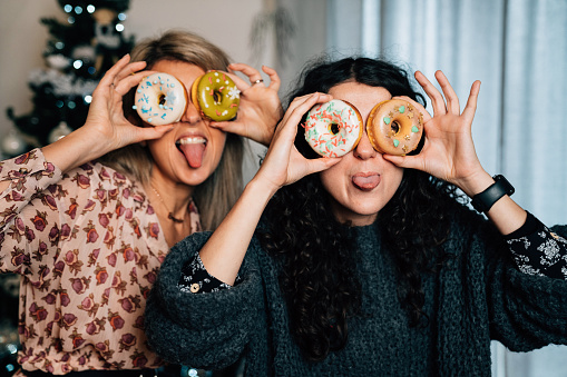 Millennial women playing together with donuts at home. Christmas tree in the background.