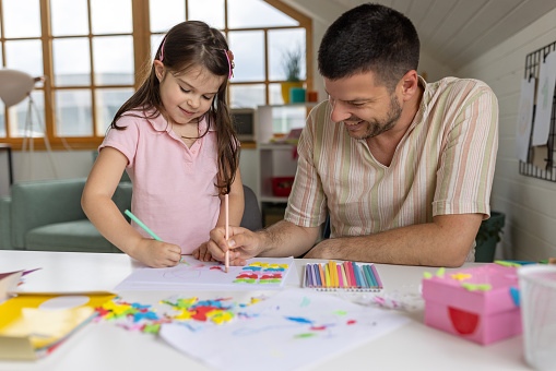 A little Caucasian girl drawing with her father in the living room of their apartment