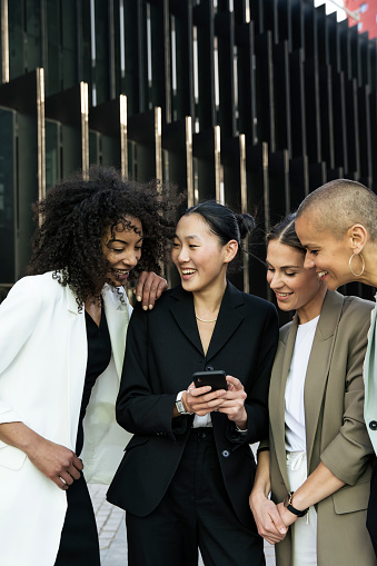 Diverse businesswomen group looking a phone and laughing standing in the street. Casual meeting between young females executives checking their smartphone outdoors.