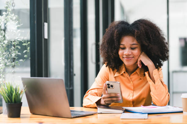 istock African businesswoman using smartphone while working on a laptop computer. Smiling African American woman using mobile phone app for playing games, shopping online, and ordering delivery in the office 1589570777