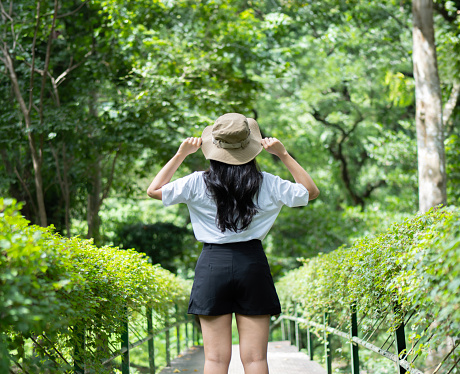 Young woman with hat hiking in forest during summer season, solo traveler walking in the forest. Travel, adventure and journey concept