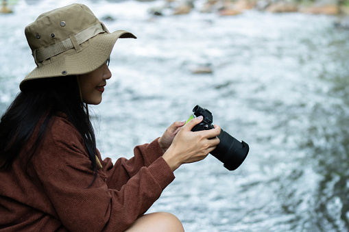 Side view of happy Asian woman with photo camera shooting tropical waterfall. Summer vacation Concept of wanderlust and adventure