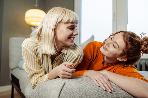 Two young Caucasian female friends are lying on a sofa and smiling while looking at each other.