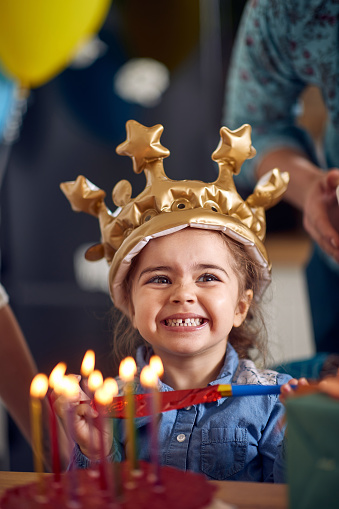 Adorable little birthday girl wearing inflatable golden crown, sitting in front of cake with lit candles, smiling feeling joyful. Birthday, celebration, family concept.