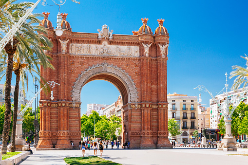 The Arc de Triomf, a famous landmark in Barcelona, Spain. The arch is made of red brick and has ornate sculptures on top, representing various historical and artistic themes. The arch is in the center of a large plaza with palm trees and lampposts. There are people walking around the plaza and a few cars on the street, indicating a lively and busy atmosphere. The sky is blue and the weather appears to be sunny and warm, suggesting a pleasant day for sightseeing. The buildings in the background are typical of Barcelona’s architecture, with colorful facades and balconies.