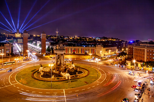 A panoramic view of Barcelona at night, with the Plaça d’Espanya and the Magic Fountain in the foreground. The fountain is illuminated by colorful lights and creates a contrast with the dark sky. The plaza is a large circular space with a fountain in the center and several roads radiating from it. The plaza is surrounded by buildings of different styles and heights, including the Museu Nacional d’Art de Catalunya, which has a classical facade and a clock tower. The museum emits blue beams of light that reach the sky, creating a spectacular effect. The image also captures the movement of the cars on the roads, creating light trails that add dynamism and depth to the scene. The image is taken from a high vantage point, allowing a wide perspective of the cityscape.