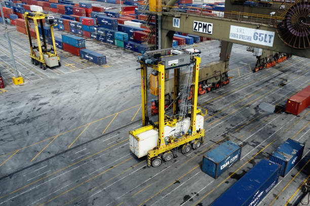 A yellow straddle carrier loaded with white reefer container in Kingston Freeport Terminal. Kingston, Jamaica - 06 07 2023: A yellow straddle carrier loaded with white reefer container in Kingston Freeport Terminal passing under the gantry crane. There are plent of other boxes from shippers. kingston harbour stock pictures, royalty-free photos & images