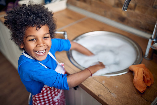 Young Afro-American boy takes on the responsibility of washing dishes with great enthusiasm. Donning a colorful apron, he stands confidently at the sink, surrounded by a sea of soapy suds.