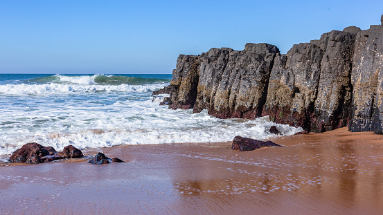 Beach black rock wall headland into sand on waters edge towards blue ocean waves and horizon blue sky landscape.