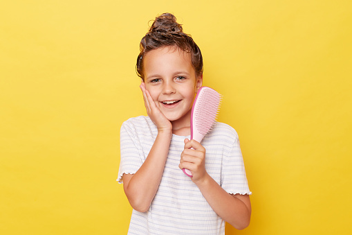 Cute smiling little girl with wet hair wearing casual white T-shirt standing isolated over yellow background doing hair care procedures holding comb