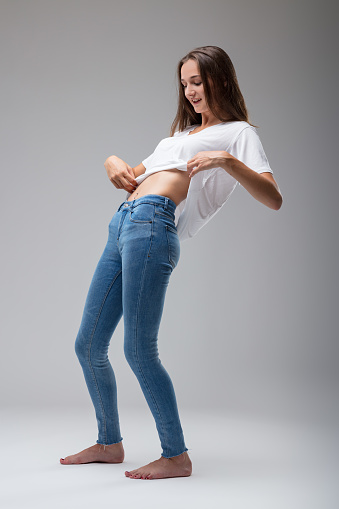 full-length portrait captures a young woman in jeans, lifting her white shirt to reveal her perfect abdomen, a sight she inspects with pride