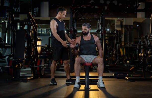 istock young trainer coaching handsome muscular man lifting dumbbell weight sitting on bench at modern gym 1589851838