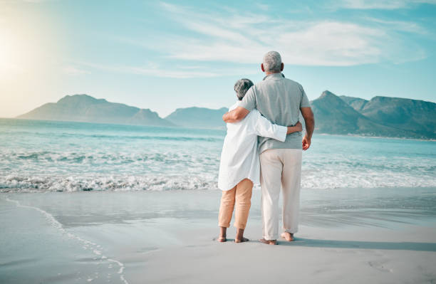 espalda, abrazo o pareja de ancianos en la playa para relajarse con amor, cuidado o apoyo en las vacaciones de verano en la naturaleza. jubilación, hombre maduro o mujer mayor en el mar o en el océano para viajar de vacaciones juntos mirando a la vista - jubilación fotografías e imágenes de stock