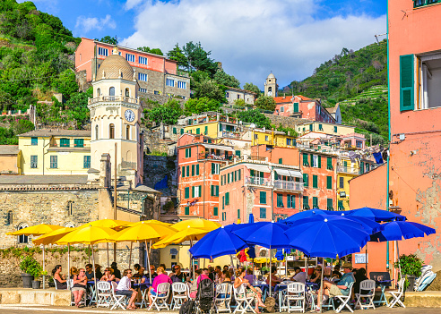 Vernazza, Cinque Terre, Liguria, Italy – May 23, 2018: Tourists sitting in restaurants in the town square of Vernazza with houses, church and villas in the background.