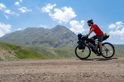 Tour bike in bikepacking order. Cyclist in red jersey with helmet. High altitude mountainous area Mediterranean region. Dirt road. Camper riding bike. Side angle. Less cloudy weather clear sunny. Steep mountainous region.