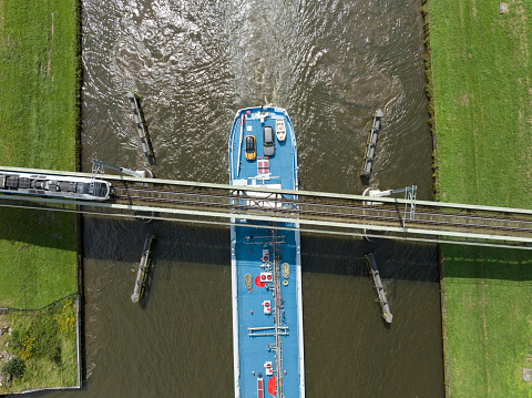 Train driving over a railway bridge crossing a canal while a ship is passing. Overhad drone view on the train bridge and bulk tanker ship.