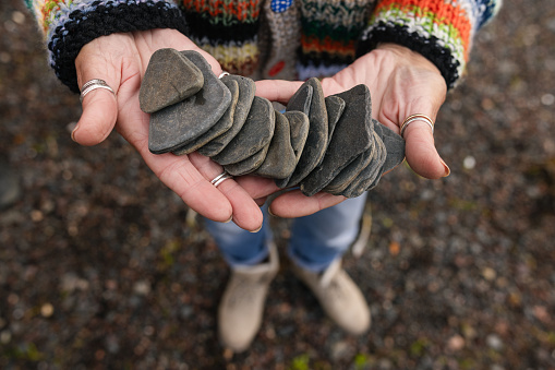 An overhead shot of a pair of hands holding a collection of flat grey stones which have been collected on a beach in Torridon, Scotland. There are several rings on the hands and the person is wearing a colourful knitted cardigan which is in partial view.