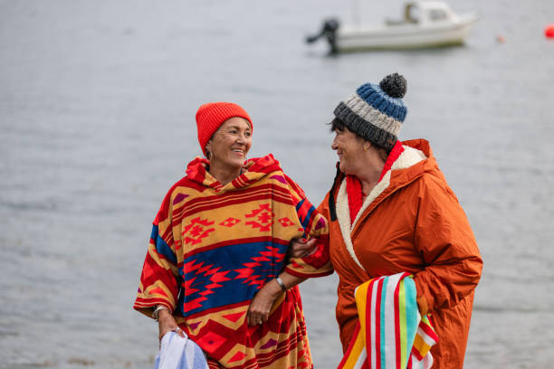 Mature Friendship at its Finest A waist-up shot of two senior female adults walking arm-in-arm along the waters edge in Torridon, Scotland. They are wearing dry robes and beanies before going for a swim in the sea loch. In the bay behind there is a boat which is in blurred focus. woman cold water swimming stock pictures, royalty-free photos & images