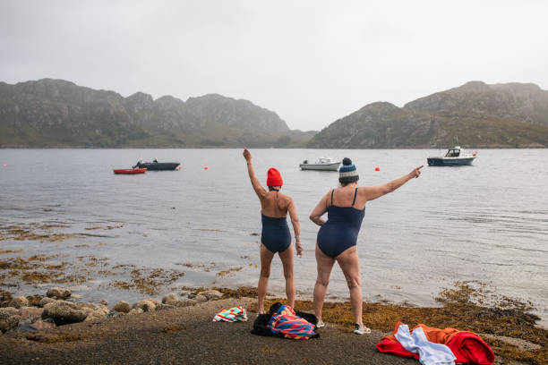 Dance Moves on the Shoreline A full-length wide shot of two senior female adults standing on the waters edge in Torridon, Scotland. They are wearing swimsuits and knitted bobble hats whilst dancing to warm up before going for a swim in the cold autumn sea. In the bay there are boats and hills in the distance. woman cold water swimming stock pictures, royalty-free photos & images