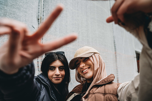 Two young adult girls are making funny gestures to the camera. One of them is wearing a fashionable hijab with an hat.