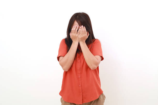 Frustrated young asian woman standing while covering her face. Isolated on white