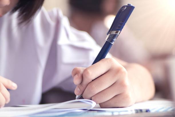 istock Close up hand of students reading and taking exam sheets exercises answer with stress in classroom 1589967095