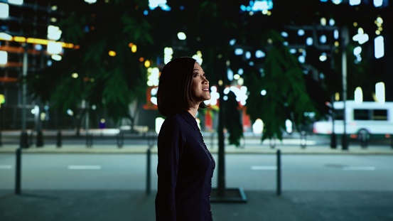Asian person strolling near city centre, looking at modern skyscrapers while she walks on sidewalk under streetlamps. Woman being alone downtown at nighttime, smiling on promenade.
