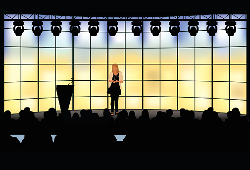 Female presenter presenting in front of silhouetted crowed.