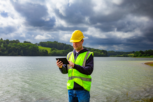 Portrait of a male maintenance engineer working outdoors at the reservoir. He is dressed in high visibility vest and yellow hardhat. He is analysing data using a digital tablet.