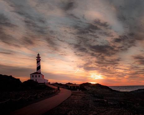 Sunrise image of lighthouse located in Menorca (Spain). Beauty sky with dramatic clouds. Orange colors and lights reflections make a nice landscape.