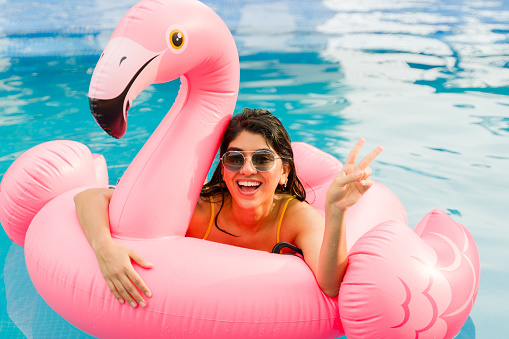 Funny latin young woman with sunglasses making the peace sign while having fun in a pink inflatable after swimming in the pool
