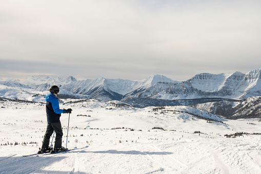 A skier is admiring the scenic mountain view in the Canadian Rocky Mountains surrounded by snowcapped mountains. It is a cloudy day and the sun is shining through the thin haze. The snow conditions are pristine, fresh powder snow fell the night before.
