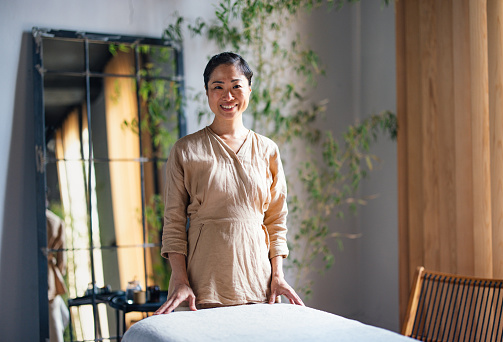 Beautiful smiling Asian masseuse standing next to the bed at the spa ready to give a massage. She is smiling and looking at the camera.