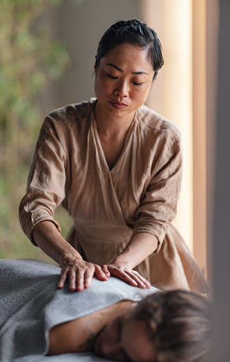 Close up shot of a beautiful Asian female masseuse giving a massage to a woman lying face down on the bed. They are in a spa center or a massage parlour. The massage therapist placing a towel over her female customer's back.