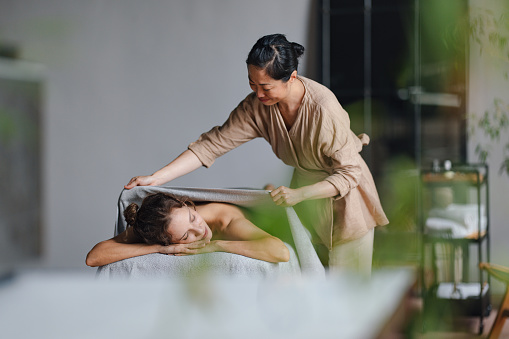 Beautiful smiling Asian masseuse standing next to a bed and placing a towel over a happy customer's back after a giving her a massage. The woman lying on the bed is relaxing with her eyes closed.
