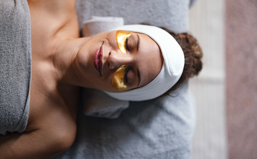 Close up shot of a beautiful woman lying on the bed at the spa centre, relaxing and enjoying having a golden eye mask. She is smiling while having her eyes closed.