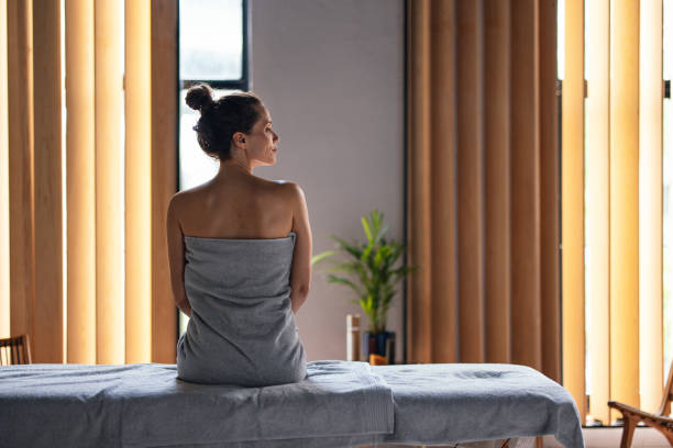 Beautiful Woman Sitting on the Massage Bed at the Spa Beautiful woman sitting on the bed waiting to get a massage or a beauty treatment. She is sitting with her back turned and looking on the side. She is wrapped in a towel. woman wrapped in bath towel stock pictures, royalty-free photos & images