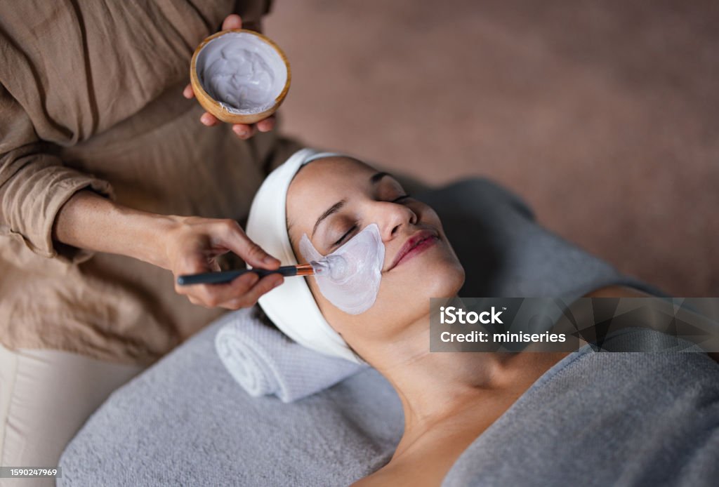 Beautiful Woman Enjoying Receiving a Facial Treatment at the Spa Close up shot of a beautiful smiling woman lying with her eyes closed while another woman is applying a face mask on her in the spa center. She is relaxed and enjoys the treatment. Facial Mask - Beauty Product Stock Photo